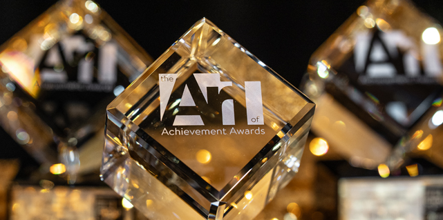 Multiple crystal cubes with the Art of Achievement Award logo etched on the front are lined up on a table covered in shimmering tablecloth